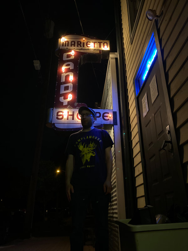 A man stands casually, hands by his side, in front of a retro Cany Shoppe sign at night