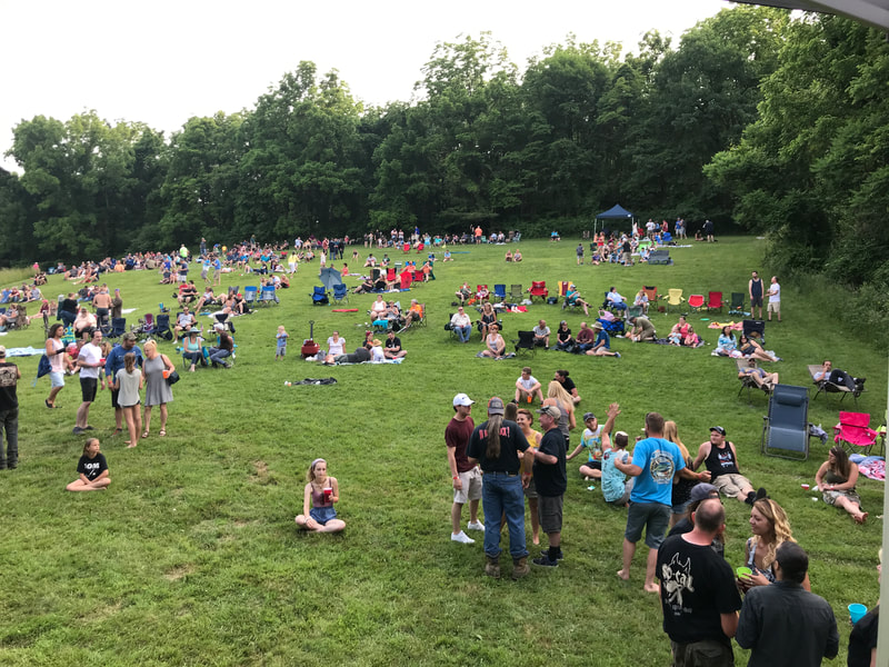 A large crowds spreads out on picnic blankets and chairs on a wide green expanse of hillside.