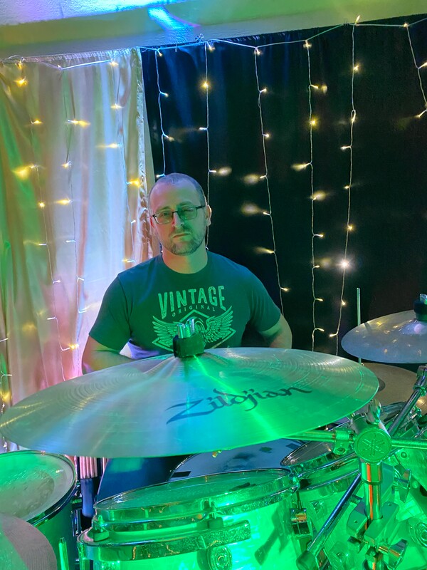 A male drummer wearing glasses and a t-shirt with the words "vintage" is seated behind his cymbals at his drum kit