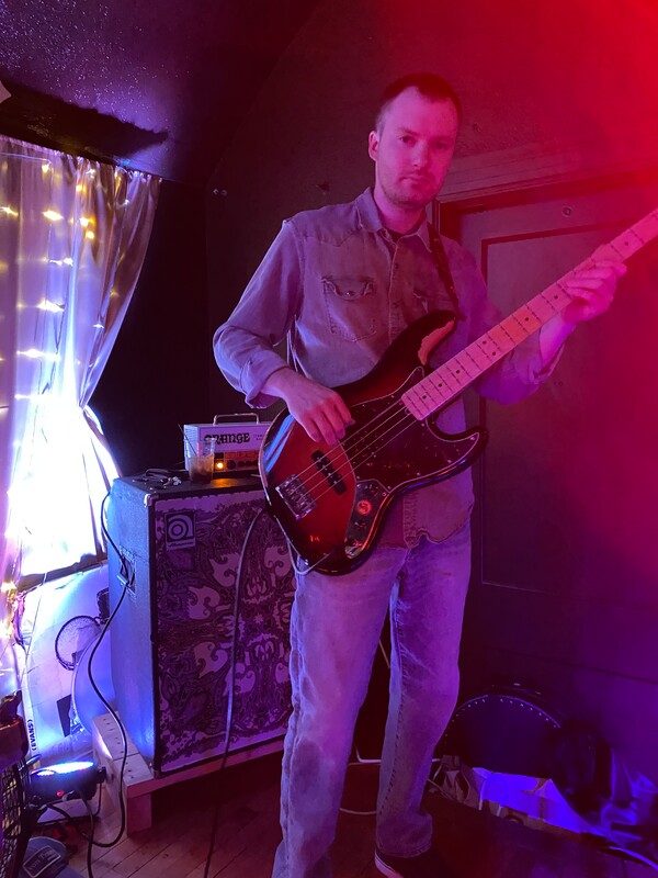 A male bass guitarist stands in the corner of a stage, playing his guitar in front of string lights and his amplifier