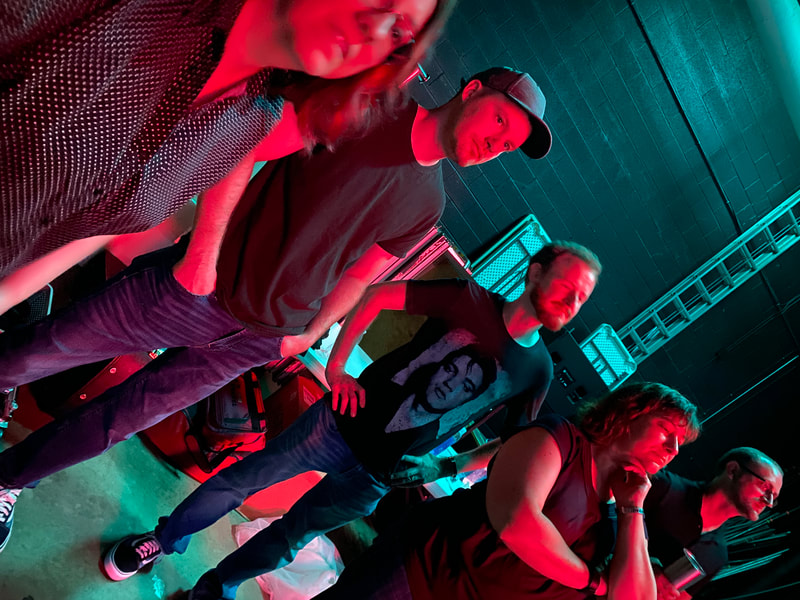 Collinsville Discount Band members stand back stage at the Appell Center in York, faces brightly illuminated in warm red color while the walls are washed in cool teal light.