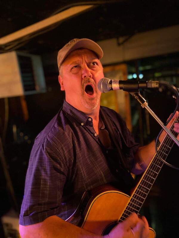 a man wearing a hat makes a silly face while singing into a microphone and holding an acoustic guitar