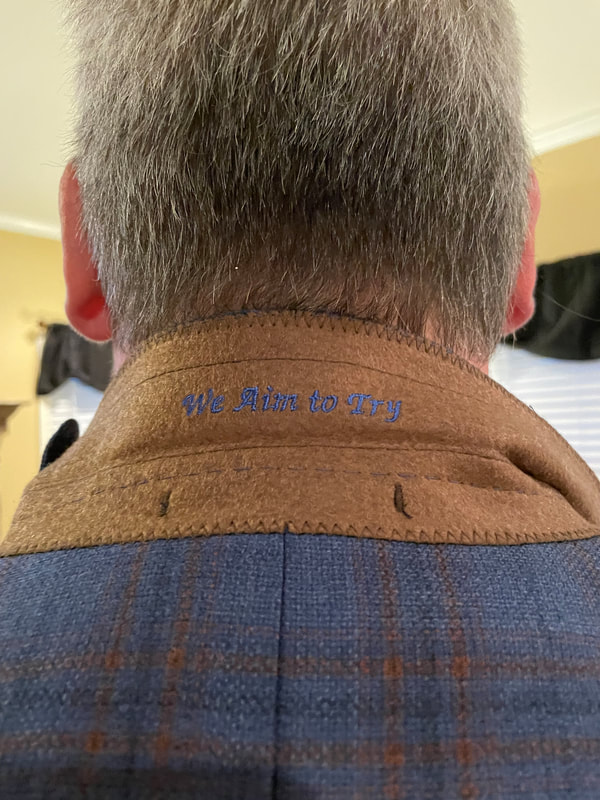 A jacket collar is flipped up to reveal the embroidered words "We Aim To Try" 