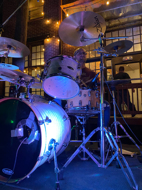 Cool purple light shines on a drum kit. A man sits on the stool behind the kit.