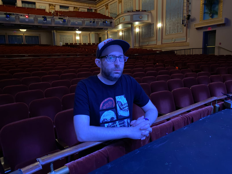 A man wearing a ballcap and glasses, leans on his elbows against the edge of a stage in a theater.