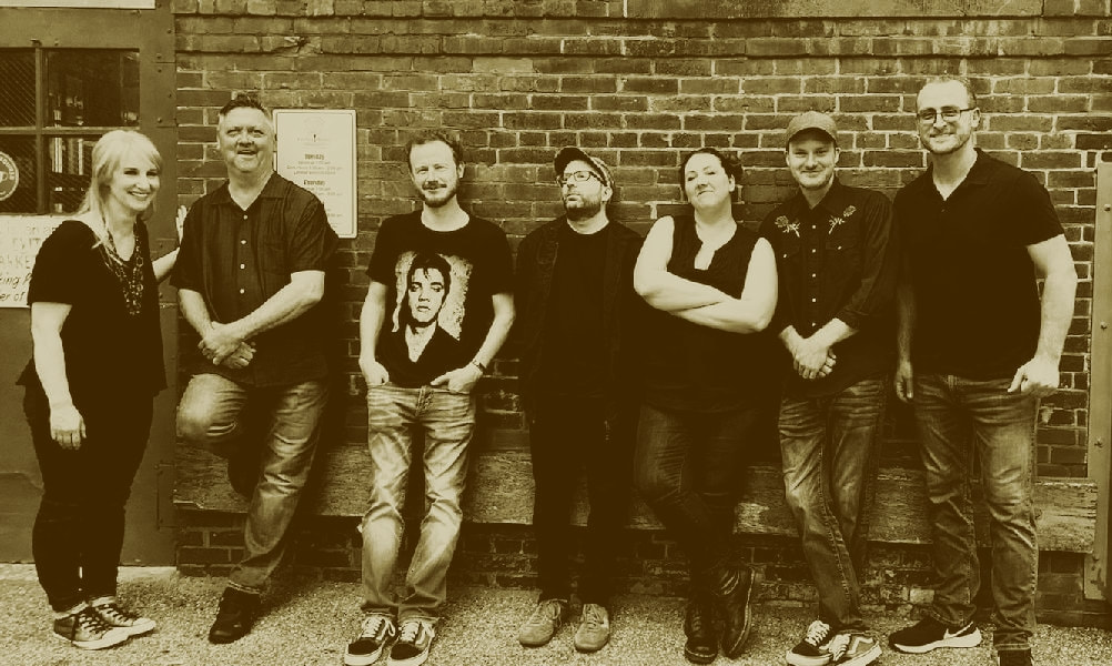 a warm toned photo of a rock band leaning casually against a brick building.