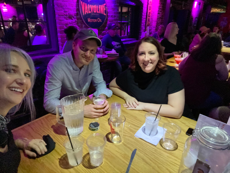 Three smiling friends sit around a bar height table full of glasses and a pitcher.