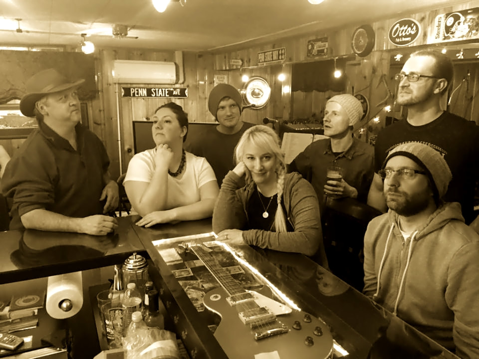 a group of seven people, two female and five males, gather casually around a glass top bar in a basement with knotty pine walls.
