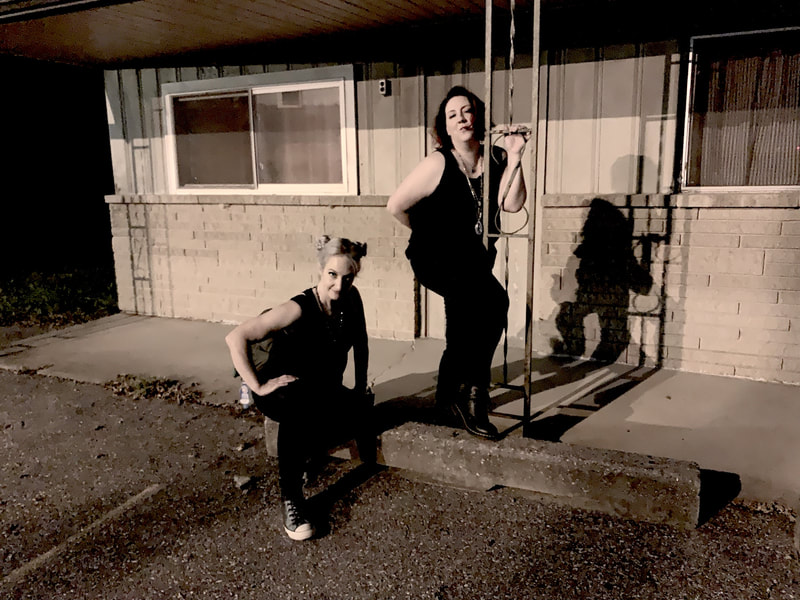 Singers Jackieraye and Jen Plonk strike a diva pose in front of a retro motel in the dim lights of a parking lot.